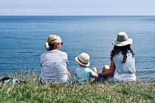 Zwei Frauen und ein Kind, alle mit Hüten, sitzen an einem Küstenabschnitt auf einer Wiese und blicken auf das Meer.