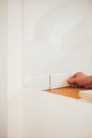 Das Bild zeigt die Hand einer Person, die über eine helle Holzoberfläche kleine Fließen montiert, mit einem Fokus auf die Handaktion gegenüber einem überwiegend weißen Hintergrund.