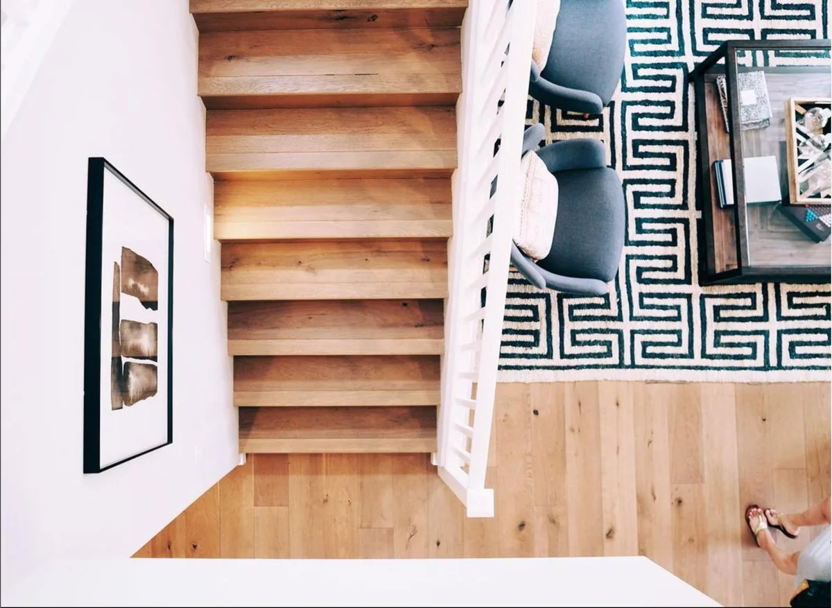 Das Bild bietet eine Vogelperspektive auf eine hölzerne Treppenkonstruktion, die in einen Raum mit stilvollem Interieur führt, inklusive eines Teppichs mit geometrischem Muster und modernen Möbeln.