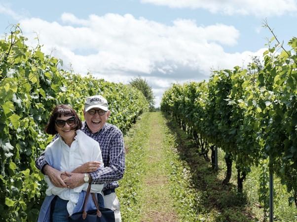 Ein älteres Paar umarmt sich liebevoll in einem Weinberg, beide lächelnd und sichtlich glücklich, umgeben von Reben und einem blauen Himmel mit weißen Wolken.