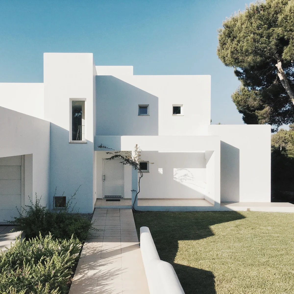 Modernes, kubistisches weißes Haus mit geometrischen Formen, umgeben von einem gepflegten Rasen, einem schlanken jungen Baum auf der Vorderseite und einem großen Kiefernbaum auf der rechten Seite unter einem klaren blauen Himmel.