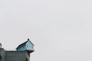 Ein kleines, auf dem Kopf stehendes blaues Haus, das an der Kante eines grauen Betongebäudes balanciert, unter einem bewölkten Himmel, der den Eindruck erweckt, als würde das Haus gleich hinunterfallen.