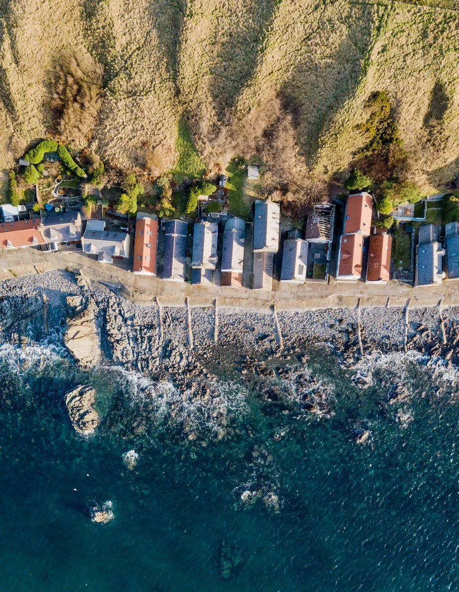 Luftaufnahme einer Küstenlinie mit einer Reihe von Häusern entlang eines Wellenbrechers, grenzend an steile grüne Hänge auf einer Seite und das offene Meer auf der anderen.