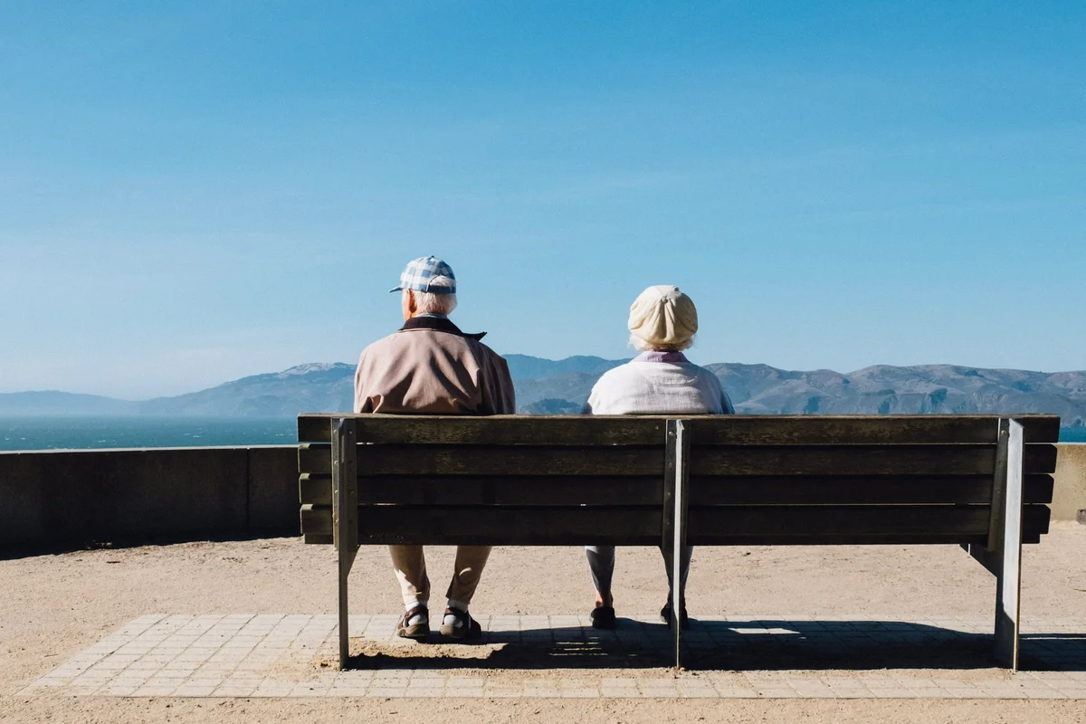 Zwei ältere Personen, die nebeneinander auf einer Parkbank sitzen und auf eine malerische Landschaft mit Bergen und klarem Himmel in der Ferne blicken.