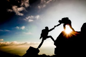Eine Silhouette einer Person, die auf einen Felsvorsprung klettert und von einer anderen Person von oben eine helfende Hand erhält, gegen einen leuchtend blauen Himmel mit der Sonne, die direkt hinter dem Fels leuchtet und einen dramatischen Lichteinfall und Schatteneffekt erzeugt. Das Bild vermittelt das Gefühl von Vertrauen.